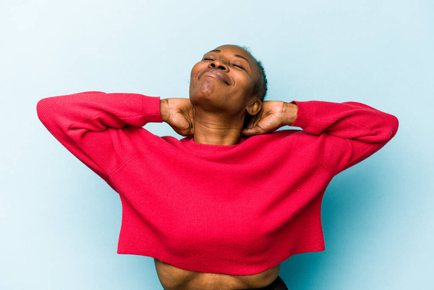 Mujer afroamericana joven aislada sobre fondo azul sintiéndose segura, con las manos detrás de la cabeza. - Foto, Imagen