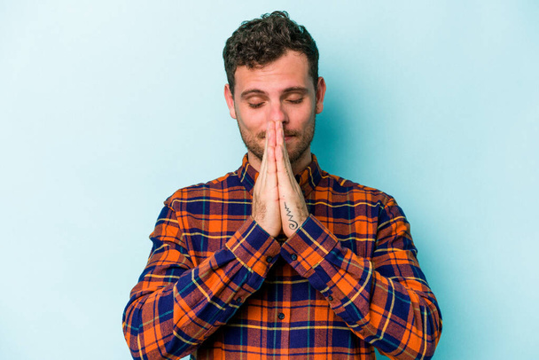 Young caucasian man isolated on blue background holding hands in pray near mouth, feels confident. - Фото, зображення