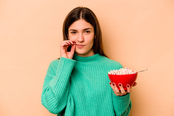 Latin kökenli genç bir kadın bej arka planda izole edilmiş mısır gevreği yiyor ve parmakları dudaklarında bir sır saklıyor.. - Fotoğraf, Görsel