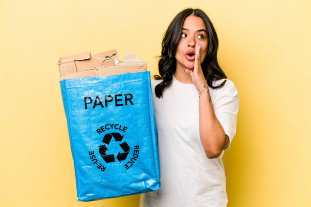 Jovem hispânica reciclando papel isolado no fundo amarelo está dizendo um segredo quente travagem notícias e olhando para o lado - Foto, Imagem