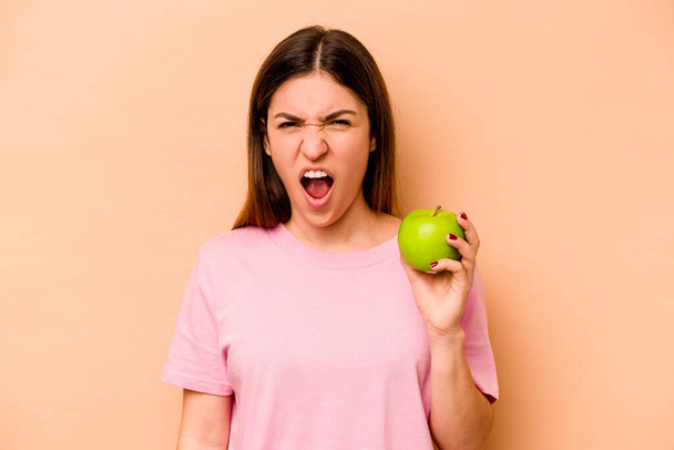 Elinde bej arka planda izole edilmiş bir elma tutan genç İspanyol kadın çok sinirli ve agresif bağırıyor.. - Fotoğraf, Görsel