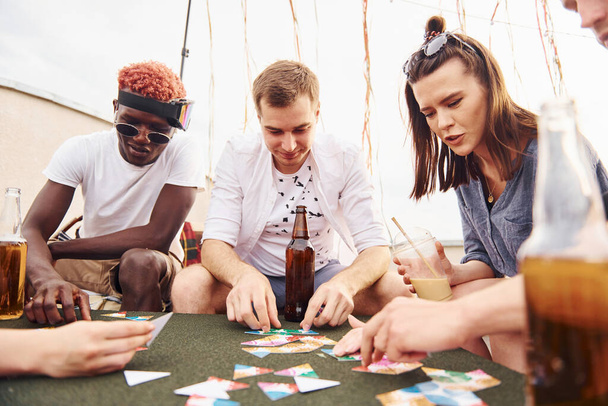 Playing card game. Group of young people in casual clothes have a party at rooftop together at daytime. - Photo, image