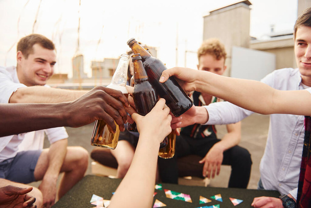 Doing cheers by bottles with beer. Group of young people in casual clothes have a party at rooftop together at daytime. - Foto, afbeelding