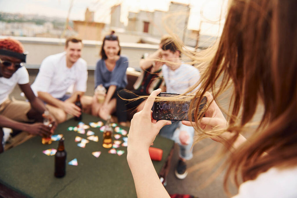 女の子がカードゲームをするときに写真を撮る。カジュアルな服装の若者のグループは、昼間一緒に屋上でパーティーを持っています. - 写真・画像