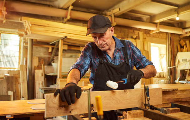 Üniformalı bir marangozun portresi marangozluk fabrikasında ahşap parmaklıkları el baskısıyla yapıştırıyor. - Fotoğraf, Görsel