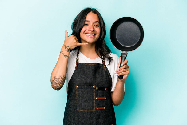 Jeune cuisinière hispanique femme tenant poêle isolée sur fond bleu montrant un geste d'appel téléphonique mobile avec les doigts. - Photo, image