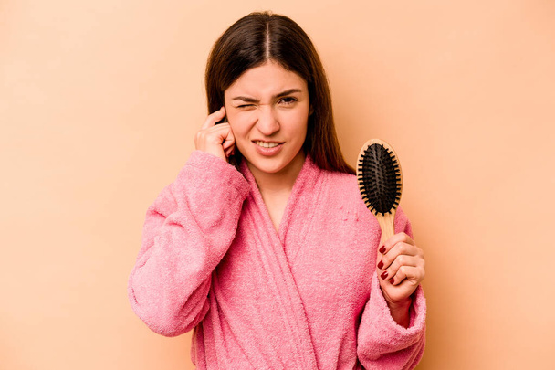 Junge hispanische Frau hält Haarbürste isoliert auf beigem Hintergrund und bedeckt Ohren mit Händen. - Foto, Bild