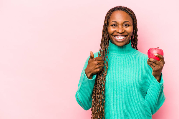 Young African American woman holding an apple isolated on pink background smiling and raising thumb up - Photo, Image