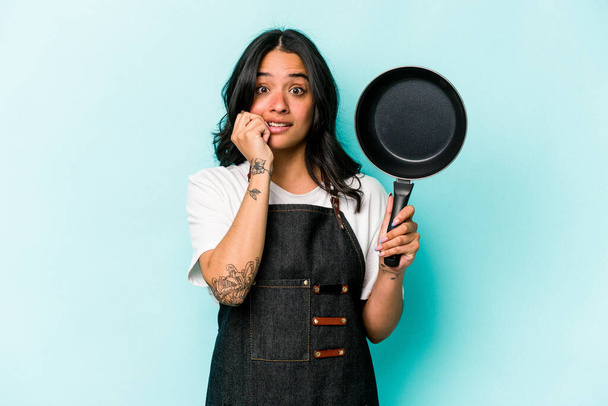 Jeune cuisinière hispanique femme tenant poêle isolée sur fond bleu ongles mordants, nerveux et très anxieux. - Photo, image