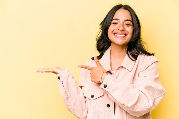 Junge hispanische Frau isoliert auf gelbem Hintergrund aufgeregt hält eine Kopie Raum auf der Handfläche. - Foto, Bild