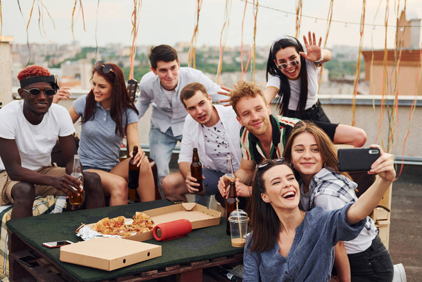 Girl making selfie. With delicious pizza. Group of young people in casual clothes have a party at rooftop together at daytime. - Photo, image