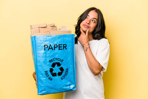 Jovem mulher hispânica reciclagem de papel isolado no fundo amarelo olhando para os lados com expressão duvidosa e cética. - Foto, Imagem