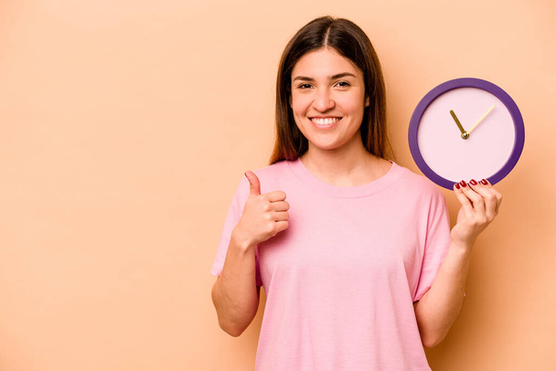 Mujer hispana joven sosteniendo un reloj aislado sobre fondo beige sonriendo y levantando el pulgar hacia arriba - Foto, imagen