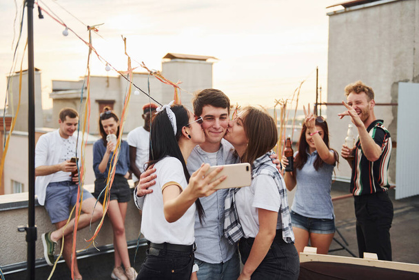 Two girls kissing one man. Taking selfie. Group of young people in casual clothes have a party at rooftop together at daytime. - Valokuva, kuva
