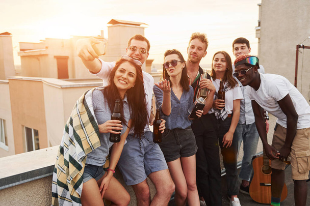 Taking selfie. Beautiful sunshine. Group of young people in casual clothes have a party at rooftop together at daytime. - Photo, Image