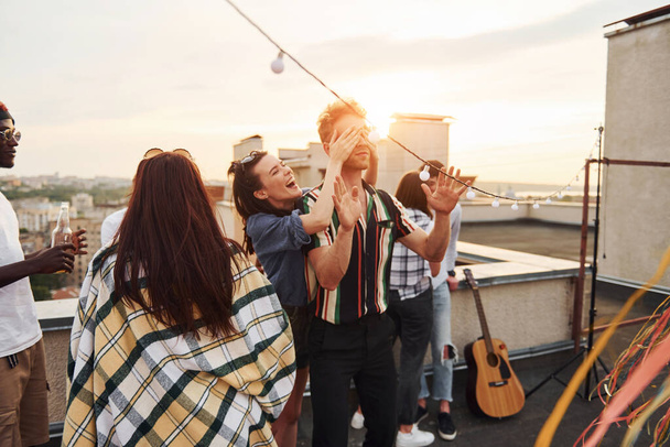 Playing game. Man's eyes covered by hands. Group of young people in casual clothes have a party at rooftop together at daytime. - Foto, Imagen