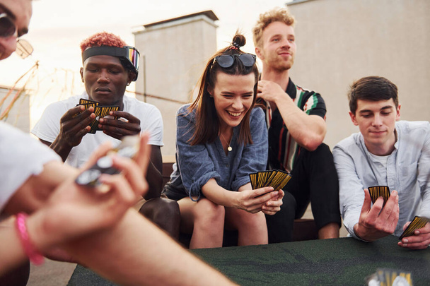 Sitting by table and playing card game. Group of young people in casual clothes have a party at rooftop together at daytime. - Fotoğraf, Görsel