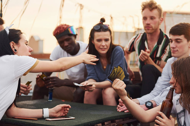 Sentado à mesa e jogando jogo de cartas. Grupo de jovens em roupas casuais fazem uma festa no telhado juntos durante o dia. - Foto, Imagem