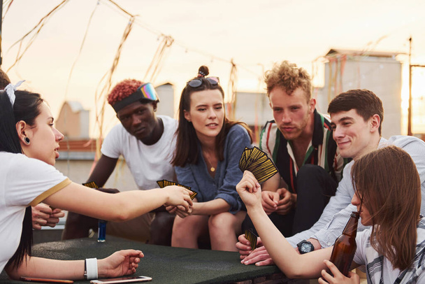 Sitting by table and playing card game. Group of young people in casual clothes have a party at rooftop together at daytime. - Fotografie, Obrázek