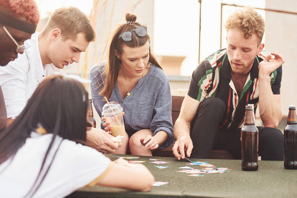 Playing game and sitting by the table. Group of young people in casual clothes have a party at rooftop together at daytime. - Fotoğraf, Görsel