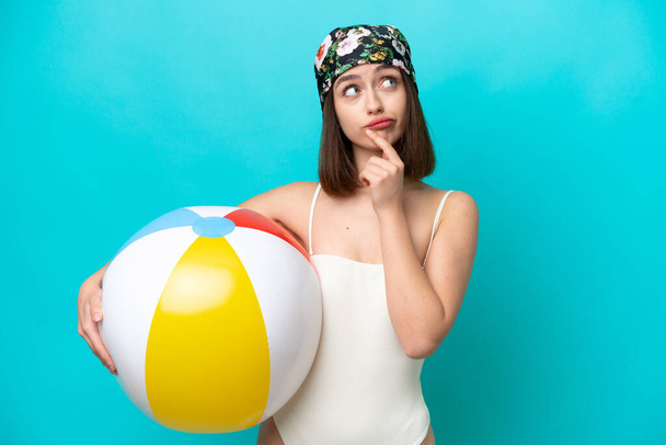 Young Ukrainian woman holding beach ball isolated on blue background having doubts - Фото, изображение