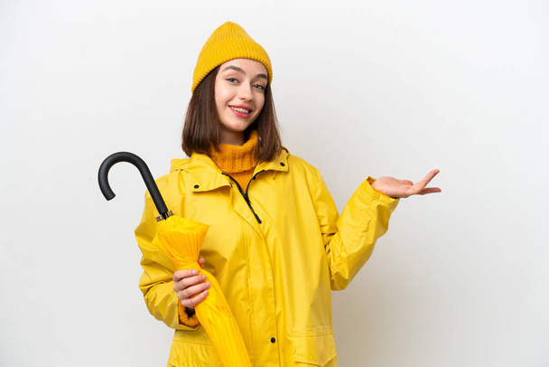 Young Ukrainian woman with rainproof coat and umbrella isolated on white background extending hands to the side for inviting to come - Foto, Imagen
