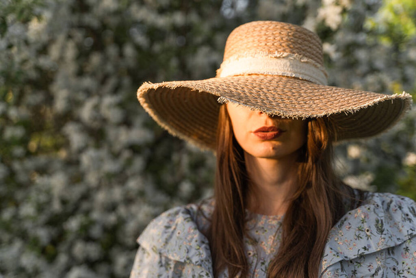 Young woman hiding her eyes under a straw hat. Vintage clothes. Walk in the spring blossom apple tree garden. Cottagecore aesthetics. Concept of female spring fashion. - Photo, image