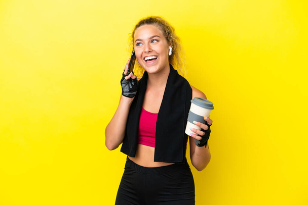 黄色の背景に隔離されたタオルを持つスポーツ女性はコーヒーを取り上げ、モバイルを保持 - 写真・画像