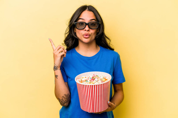 Young hispanic woman holding popcorn isolated on yellow background having some great idea, concept of creativity. - Zdjęcie, obraz