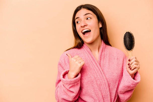 Young hispanic woman holding hairbrush isolated on beige background points with thumb finger away, laughing and carefree. - Photo, Image
