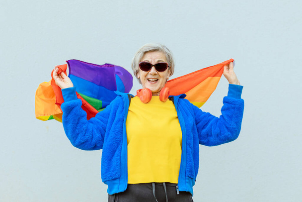 happy 80 years old woman dressed colorfully in sunglasses proudly waving the rainbow flag of the gay community - Фото, зображення