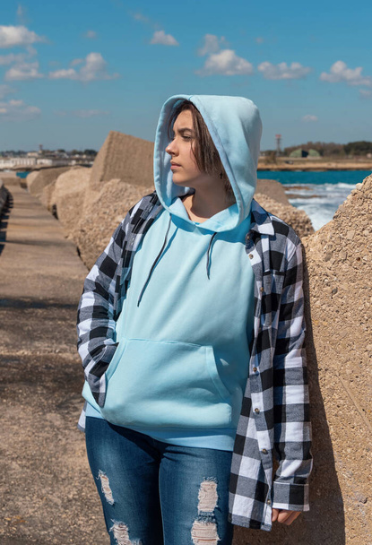 Teenager in light blue hoodie and plaid shirt looking aside. Teen girl stays outdoor near the sea, concrete blocks and blue sky, windy day. Clothing mockup, copy space - Photo, Image
