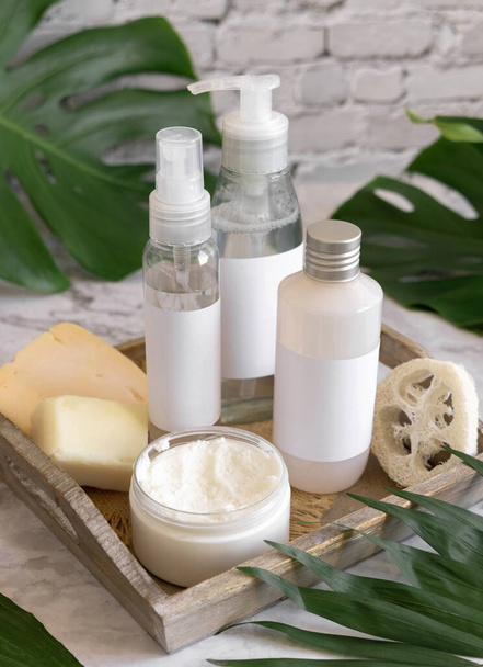 Cosmetic bottles and skin care accessories near green monstera leaves close up against white brick wall. Brand packaging mockup. Organic beauty products. Natural homemade cosmetic - Photo, Image