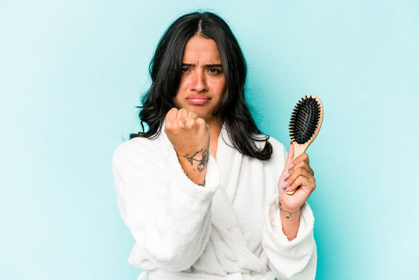 Fiatal spanyol nő gazdaság hajkefe elszigetelt kék háttér mutatja ököllel a kamera, agresszív arckifejezés. - Fotó, kép