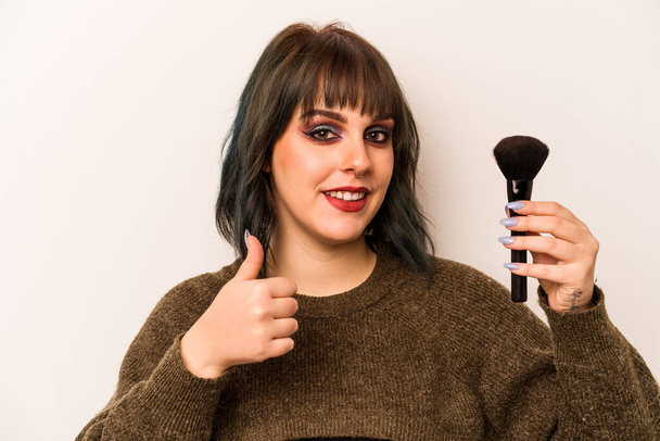 Jovem artista de maquiagem caucasiana segurando uma escova de maquiagem isolada no fundo branco sorrindo e levantando o polegar - Foto, Imagem