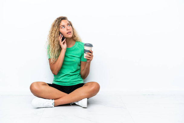 Dziewczyna z kręconymi włosami siedzi na podłodze trzymając kawę do zabrania i telefon - Zdjęcie, obraz