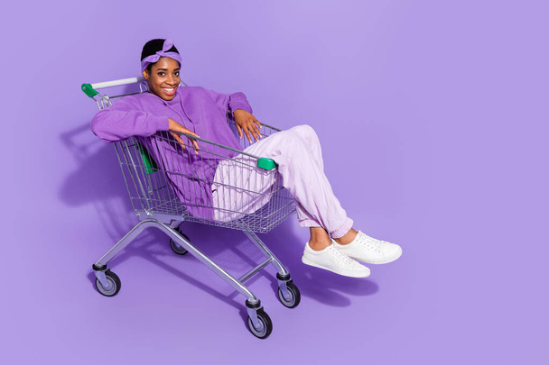 Full length photo of positive bisexual man in trendy hoodie go shopping sit pushcart isolated on violet color background - Foto, afbeelding