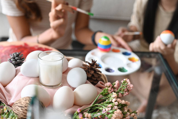 Cesta de mimbre llena de huevos y flores en la mesa en la sala de estar con la madre y la hija pintando huevo de Pascua en el fondo. - Foto, imagen