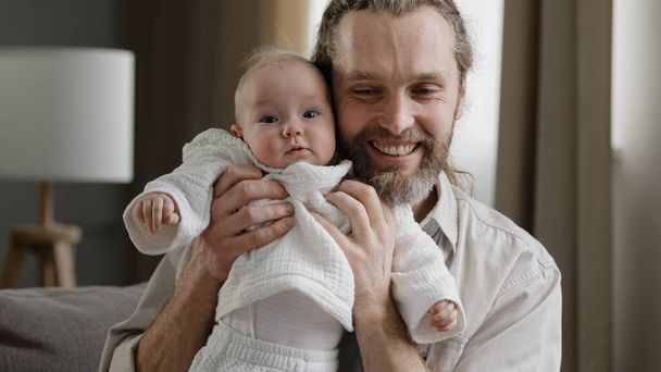 Kochający dorosły brodaty ojciec trzymający dziecko noworodka opiekuńczy tata bawiący się z małą córką syn skaczący śmiejący się mężczyzna całujący dziecko w głowę razem bawić się w domu siedząc na kanapie szczęśliwy czas rodzinny - Zdjęcie, obraz
