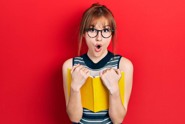Κοκκινομάλλα νεαρή γυναίκα που διαβάζει ένα βιβλίο φορώντας γυαλιά φοβισμένος και σοκαρισμένος από την έκπληξη και κατάπληκτος έκφραση, το φόβο και ενθουσιασμένος πρόσωπο.  - Φωτογραφία, εικόνα