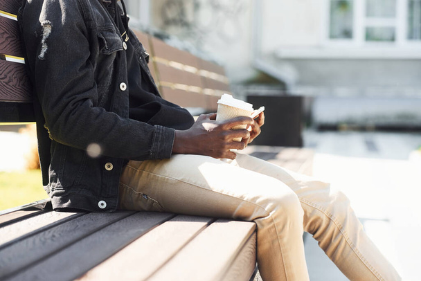 With phone and drink. Close up view of african american man in black jacket outdoors in the city sitting on bench. - Zdjęcie, obraz