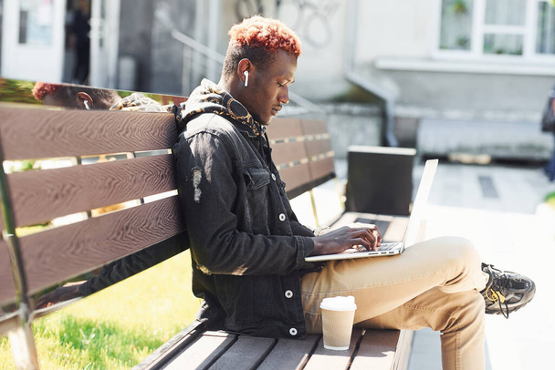 Working by using laptop. Young african american man in black jacket outdoors in the city sitting on bench. - Fotoğraf, Görsel