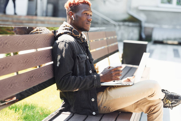 Working by using laptop. Young african american man in black jacket outdoors in the city sitting on bench. - Foto, imagen