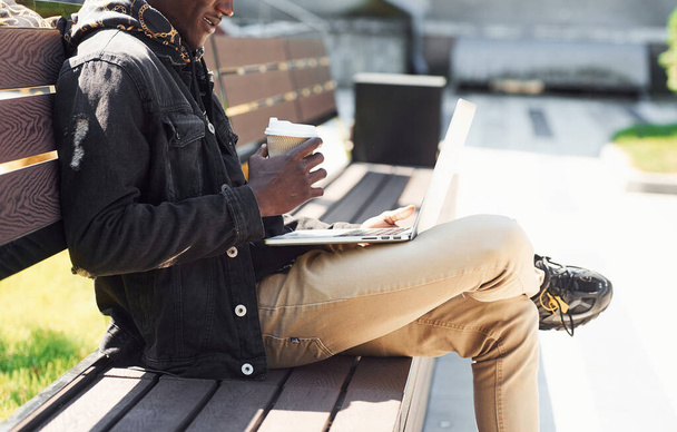Working by using laptop. Close up view of african american man in black jacket outdoors in the city sitting on bench. - Photo, Image