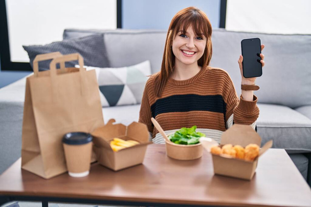 Young beautiful woman eating delivery food at the living room looking positive and happy standing and smiling with a confident smile showing teeth  - Foto, afbeelding