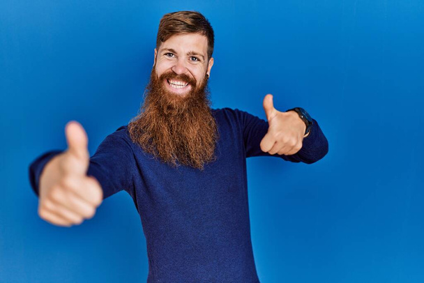 Рыжий мужчина с длинной бородой носит случайный синий свитер на синем фоне, одобряя позитивный жест рукой, большой палец вверх улыбается и счастлив за успех. жест победителя.  - Фото, изображение