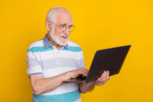 Portrait of attractive cheerful focused grey haired man using laptop writing email isolated over bright yellow color background - 写真・画像