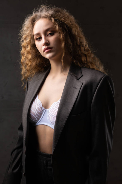 Portrait of a young red-haired curly sexy girl in a jacket with long hair in the studio. Shows off her curvaceous breasts in a bra. - Foto, immagini