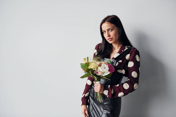 Beautiful young brunette in black skirt standing indoors against white background with bouquet in hands. - Valokuva, kuva