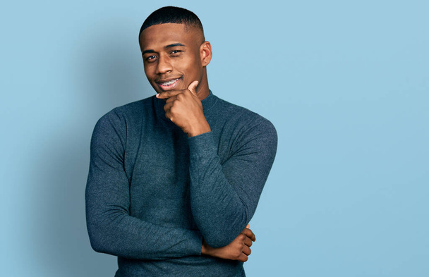 Ein junger schwarzer Mann in lässigem Pullover lächelt selbstbewusst in die Kamera, die Arme verschränkt und die Hand am Kinn. Positives Denken.  - Foto, Bild